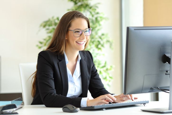 Businesswoman wearing glasses working with a desktop computer at office checking automated emails and smiling