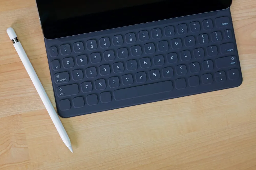 An iPad case with built-in keyboard, along with a stylus, sit on a wooden desk.